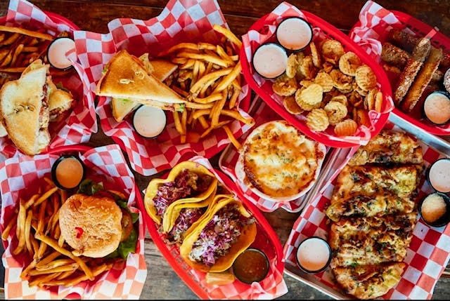 Spread of food at The Roost Lewisville — burgers, tacos, fries, fried pickles and queso in red baskets